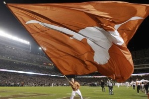 A male cheerleader from UT is waving a gigantic burnt orange flag with a white longhorn head symbol on it. He is wearing an orange shirt and white pants. He is standing on the football field. It is night time.